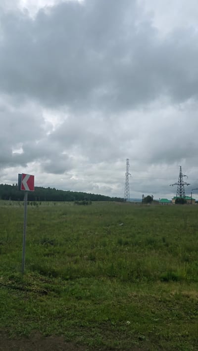 The image, captured in the vicinity of Актюбинский, Russia, presents an expansive outdoor scene under an overcast, uniformly grey sky with dense cloud cover. The foreground consists of a dark earthen strip at the bottom transitioning into an extensive field of green grass, characterized by varied textures and growth. On the far left, a grey metal post supports a partially visible square road sign; its face, angled towards the viewer, shows a white background with a prominent red chevron pointing left, indicating a sharp turn. The reverse side of the sign is dark grey or black. In the mid-ground, a dense line of dark green trees, forming a distant treeline or forest, spans the horizon from the left to the right. Two distinct lattice-structure electricity transmission pylons are visible on the right side of the frame. The taller, slender pylon is centrally located, extending high into the sky, while a second, broader pylon stands further to the right. Multiple power lines stretch horizontally across the frame, connecting these pylons and receding into the distance. Behind and to the right of the broader pylon, a cluster of buildings is partially visible, featuring prominent green roofs and light-colored walls. No individuals or animals are present in the scene.