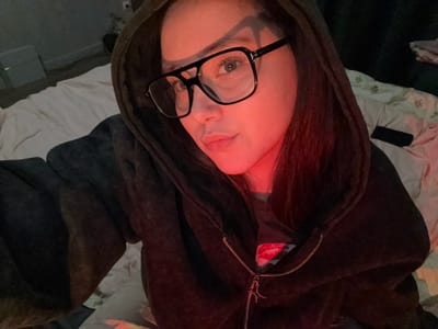 A female individual is depicted in an indoor, dimly lit setting, situated on what appears to be a bed in the city of Prikubanskii, Russia. She is shown in a mid-shot, looking directly towards the camera. She has long, dark hair, partially covered by a dark hooded sweatshirt or jacket. She wears large, black-framed eyeglasses. Her face exhibits a neutral expression, and a strong red light illuminates her right cheek, right eye, and the right side of her hair (viewer's left side). The garment she is wearing is a dark, possibly charcoal or black, zippered hoodie, which appears to be made of a soft, textured material. The metallic zipper pull has a small, star-shaped charm attached. Underneath the hoodie, a dark t-shirt with a red lip graphic is partially visible. The background on the viewer's left includes a light-colored duvet or comforter, and further back, a dark wall with a white rectangular object and a wall fixture. On the viewer's right, a section of a light-colored, possibly patterned comforter is visible, along with a dark, rectangular object resembling a smartphone or remote control lying on the bed. The overall lighting conditions are low, with the prominent red light source creating distinct shadows and highlights.