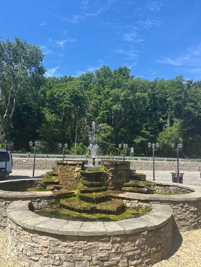 A tiered stone fountain with moss-covered ledges is the central focus of this outdoor scene. Water cascades down the levels of the fountain, creating a gentle spray. The fountain is surrounded by a circular stone wall, and the ground is covered in small, light-colored gravel.
Several decorative lampposts are positioned around the fountain. To the left, the rear of a white van is visible, parked on a paved area. In the background, a low stone wall runs horizontally, separating the fountain area from a road or pathway. Beyond that, a dense forest of green trees forms a natural backdrop under a bright blue sky with scattered white clouds.
The image appears to be taken on a sunny day, with clear visibility. The overall setting suggests a park, resort, or public space. The location provided is Shirokaya Schel, Russia, however, there are no distinct visual elements in the image that confirm this specific location.