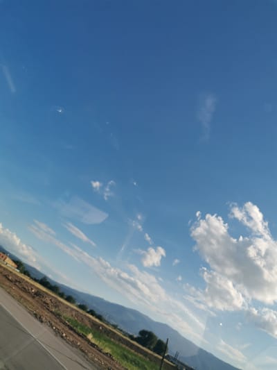 The image is a tilted photograph of a clear blue sky with scattered white clouds. In the foreground, a diagonal asphalt road runs from the bottom left corner upwards and to the right. Adjacent to the road is a dirt embankment, some green grass, and further back, a line of trees. In the distance, hazy mountains form the horizon. A building with an orange roof is visible on the far left, partially obscured by the embankment. The scene appears to be captured from a moving vehicle, as evidenced by reflections on a glass surface, including what looks like a small, translucent object with text and a barcode. The lighting suggests it is daytime. The location is identified as Krivina, Bulgaria, though this is not visually evident in the photograph itself.