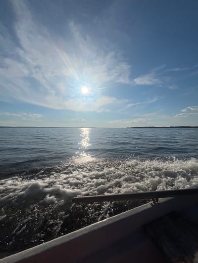 This image depicts a serene scene of a body of water, likely a river or lake, from the perspective of a moving boat. The sun shines brightly in the sky, casting a shimmering reflection on the water's surface. The sky is a clear blue, dotted with wispy white clouds. In the foreground, the wake of the boat creates a frothy trail of white foam against the dark water. The horizon is a thin line separating the sky and water, with a subtle indication of land in the distance. A portion of the boat's railing is visible on the right side of the frame, adding to the sense of movement and perspective. The overall mood is peaceful and tranquil, with the bright sunlight suggesting a clear, pleasant day. The location is identified as Siukterka, Russia.