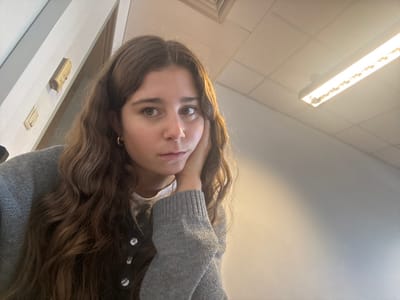 The image displays a young adult female situated indoors in what is identified as the city of Porto Salvo, Portugal. The subject has long, wavy brown hair and a light complexion, looking directly at the camera with a neutral expression. She wears a light grey knit cardigan with white buttons over a white top, a small gold hoop earring in her left ear, and a thin gold necklace. Her left hand rests against the left side of her face. The background on the left reveals a section of a light-colored wall or door frame with two light-toned rectangular objects mounted vertically. The upper right background features a white grid drop ceiling and a brightly lit rectangular fluorescent fixture emitting warm-toned light. The wall beneath the ceiling is a plain, light grey color. The scene is illuminated by artificial overhead lighting.