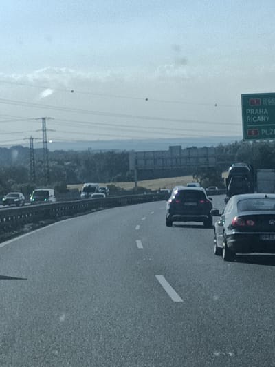 The image captures a view from a vehicle traveling on a highway. The scene depicts a multi-lane road with several cars and vans in motion. In the foreground, a dark-colored car with its taillights illuminated is visible. Further down the road, a dark SUV and other vehicles can be seen. The highway is bordered by a metal guardrail and a wooded area in the distance. Overhead power lines and pylons are also present. The sky is bright and overcast, suggesting it is daytime. A green highway sign with white lettering indicates "1 E 50" and lists destinations as "PRAHA" and "ŘÍČANY" on the top section, and "0 PLZEŇ" on the bottom section. The overall setting appears to be a highway in Všechromy, Czechia, during daylight hours with cloudy weather conditions.