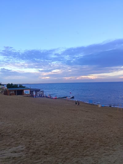 This media file depicts a beach scene at dusk. The sky is a vibrant mix of blue and purple hues, with scattered clouds tinged with the soft glow of the setting sun. The sea stretches out to the horizon, its surface rippling gently.
In the foreground, a sandy beach is visible, with footprints marking the sand. A few people are scattered across the beach, some near the water's edge and others closer to buildings. Two individuals are walking along the shore.
Along the beachfront, there are several structures. One building appears to be a small cafe or shop, with signage that includes the Russian word "МАССАЖ" (massage) and other text. Another structure, larger and darker, has text in Russian that reads "Бани На Рапе" (Baths on Rapae), suggesting it might be a bathhouse. Near this building, there are a few people gathered, and a paddleboard leans against it.
Further down the beach, a small boat is anchored in the water, flying a Russian flag. Other objects on the beach include blue chairs and what appear to be buoys. The overall atmosphere suggests a relaxed, end-of-day scene at a coastal resort. The lighting indicates it is either early morning or late evening. The presence of beach structures, people enjoying the waterfront, and the calm sea point to a recreational location.