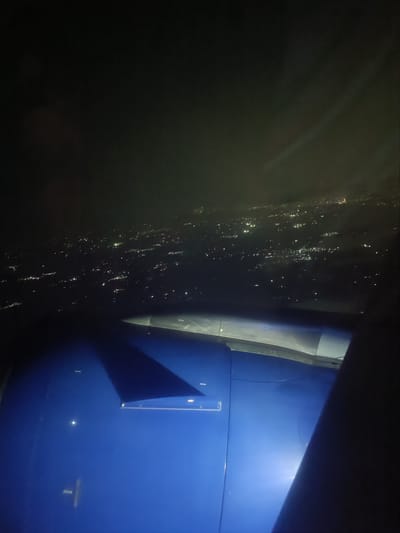 This is an aerial view of the city of Agassaim, India at night, as seen from an airplane window. The scene is dominated by the dark sky, with faint lights of the city twinkling below. Part of the airplane's wing and fuselage is visible in the foreground, painted a vibrant blue. The wing casts a shadow on the fuselage. The city lights appear as a dense pattern of small, white and yellow dots scattered across the dark landscape. Some areas of the city appear brighter, suggesting clusters of buildings or activity. The image captures the vastness of the urban sprawl seen from a high altitude.
