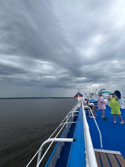 A group of people are on the deck of a ferry boat sailing on a river under a cloudy sky. The boat is moving away from the viewer, with the port side deck visible. The deck is painted blue, and there are white railings. In the foreground, a young boy in a bright green t-shirt and shorts is looking through binoculars. Next to him, a woman in a pink dress stands with her back to the camera. Further back, two men are seated on a bench. The river is dark and choppy, and a distant shoreline with trees and buildings is visible under a dramatic, overcast sky. A Russian flag is visible on the boat. The scene appears to be in the city of Ikskoe Ust'e, Russia, based on the provided context.