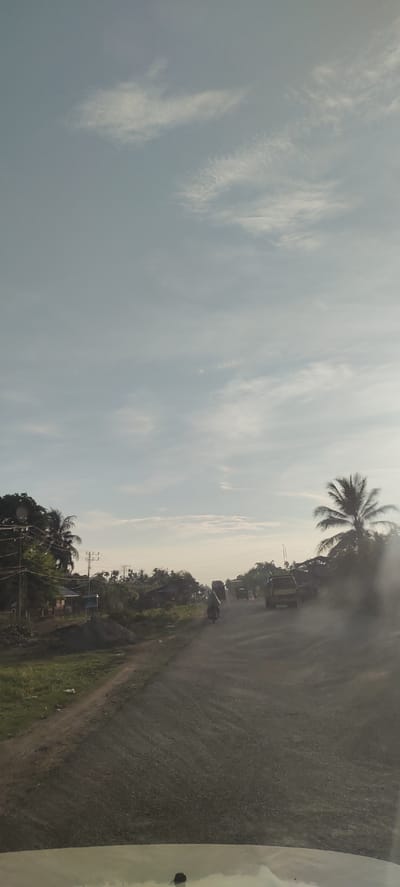 This is a photograph taken from inside a vehicle, looking out at a road in Meunasahmesjid, Indonesia. The road appears to be unpaved or gravel, and there is a noticeable amount of dust being kicked up by vehicles. On the left side of the road, there is a grassy embankment with a few buildings and power lines visible. Further down the road, a line of vehicles, including trucks and a motorcycle, are driving. Palm trees and other tropical vegetation are scattered throughout the scene. The sky is partly cloudy, with a hazy, pale blue appearance, suggesting it may be early morning or late afternoon.