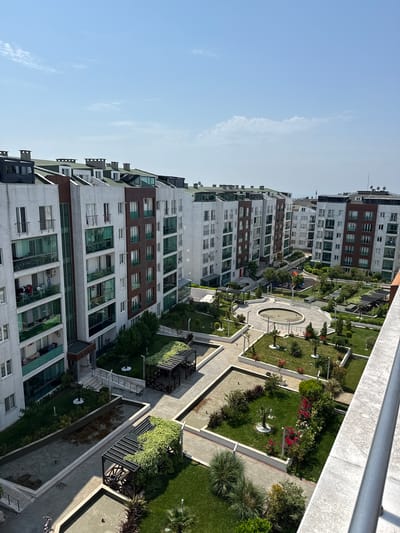 This media file presents a high-angle view of a modern residential complex in Beylikdüzü, Turkey, under a clear, bright blue sky with a few wispy clouds. The image captures several multi-story apartment buildings with contemporary architecture, featuring predominantly grey and white facades accented with dark brown panels and large glass windows and balconies. Many windows have security grates, and air conditioning units and satellite dishes are visible on the building exteriors and rooftops.
Between the buildings lies an expansive, meticulously landscaped communal garden area. This green space incorporates neatly trimmed lawns, a variety of small trees, bushes, and flowering plants, including vibrant red and white blooms. Paved pathways with geometric designs meander through the garden, connecting different sections. Notable features of the courtyard include a circular decorative fountain or water basin at the center of a paved area, and several long, rectangular water features or ponds with dark water. Wooden pergolas or trellises draped with greenery provide shaded areas, and benches are placed along the paths. In the background, a colorful children's playground with swings and a slide is visible.
The scene appears tranquil and devoid of any specific ongoing activity or event, though a bicycle is partially visible on a paved path. No people are clearly discernible in the image. The sunlight casts distinct shadows, indicating a bright daytime setting, likely morning or afternoon. No visible text is present in the image. The overall impression is one of a well-maintained, modern urban residential environment.