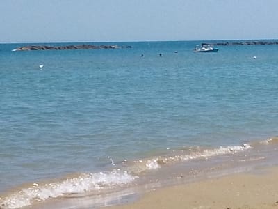 This is a wide shot of a beach on a sunny day. The ocean is calm with small waves lapping at the sandy shore. In the distance, there are two breakwaters made of large rocks. A small white boat with a blue trim is visible in the middle of the water, and a few people are swimming or floating nearby. The sky is a clear, light blue, suggesting fair weather. The image appears to be taken from the beach, looking out towards the sea. The time of day is likely midday or early afternoon due to the bright sunlight. There is no visible text in the image.