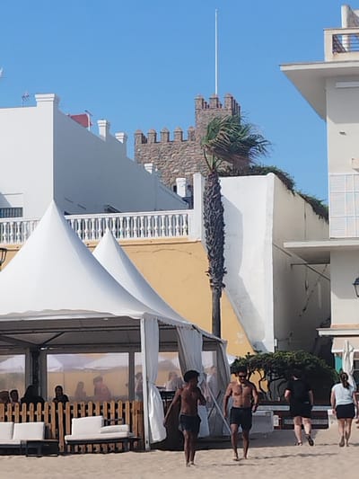 The image depicts a scene on a beach in Rota, Spain. In the foreground, two white conical tents are set up, likely for a beachside establishment. Several people are visible on the sandy beach. Two men in swim trunks are walking towards the viewer, while a couple walks away. Behind the tents, a brown wooden fence encloses seating areas with white cushions. Further in the background, a historic stone tower with crenellations stands against a clear blue sky. A tall, thin palm tree with dried fronds is situated near the tower. The buildings in the background are white, with some featuring balconies and windows. The overall atmosphere is a sunny day at the beach.