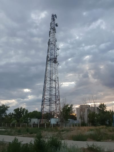 A tall, lattice communications tower dominates the frame, angled slightly to the right, with multiple antennas and dishes attached. The tower is set against a heavily clouded sky, with patches of sunlight breaking through in the upper left and center. In the middle ground, to the right of the tower, is a low-rise, utilitarian building with windows and what appears to be graffiti or markings on its facade. The building is surrounded by trees and vegetation. A wooden fence runs horizontally across the image in front of the building and some smaller utility structures. The foreground consists of a paved pathway and overgrown grass and bushes. The scene appears to be an industrial or semi-urban area, possibly on the outskirts of the city of Kurchatov, Kazakhstan, given the context of the request. There are no people visible in the image.