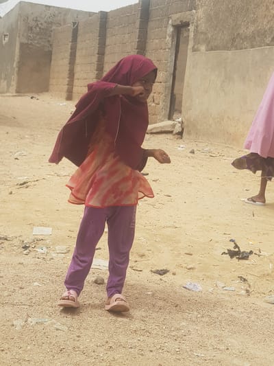 A young girl in a maroon hijab, orange patterned tunic, and purple pants stands on a sandy, littered ground in Dutsinma A, Nigeria. A building is behind her, and another person is partially visible at the top of the image. The girl's hand is raised towards her face.