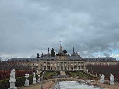 The image displays an exterior view of the Palacio Real de La Granja de San Ildefonso, situated in Real Sitio de San Ildefonso, Spain. The foreground reveals a section of the palace gardens under renovation or construction, characterized by exposed earth, scattered construction materials including white covers, and what appear to be unfinished or protected steps. Several white classical statues, some partially covered, stand on pedestals within this immediate area. Ascending towards the palace, the midground features formal, terraced gardens with manicured green hedges, bare deciduous trees, and additional white classical statues positioned along pathways and garden beds. The background is dominated by the palace structure, a multi-story Baroque building constructed of light-colored stone, featuring a symmetrical facade with numerous windows, a prominent central section adorned with columns, and a dark slate roof system punctuated by multiple spires, cupolas, and dormer windows. The sky is predominantly overcast with heavy grey clouds, though some patches of lighter sky are visible towards the upper right. No individuals are present in the visible scene.