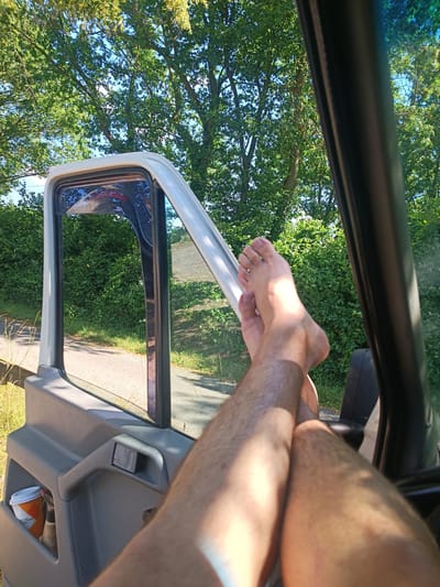 The image shows a person's bare legs and feet propped up on the open driver's side door of a vehicle. The person is inside the vehicle, with their legs extended out the open window. The setting is outdoors, with lush green trees and bushes visible in the background, suggesting a warm and sunny day. Sunlight is casting shadows on the person's legs, indicating it is likely midday. The vehicle appears to be parked on the side of a road or a path, with a glimpse of the road and grass visible outside. A coffee cup holder with a disposable cup is visible on the door panel inside the vehicle. The overall mood is relaxed and casual, with the person seemingly enjoying a moment of rest. The location is identified as Nordkirchen, Germany.