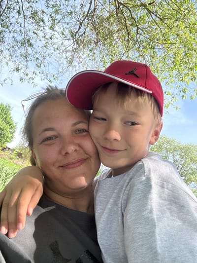 A selfie-style photograph captures a woman and a young boy outdoors, likely in the Синьково, Russia area, suggested by the presence of lush vegetation, trees with green leaves, and clear sky. The woman is in the foreground, with the boy leaning on her. The boy is wearing a red baseball cap with a black logo. Both are smiling, indicating a friendly and happy interaction. The background features branches of trees against a sky with little visible cloud cover.