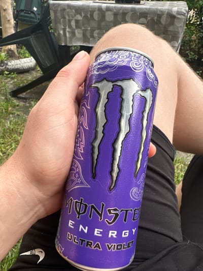 The media file shows a person's arm and hand holding a purple can of Monster Energy drink, specifically the "Ultra Violet" flavor. The can features the iconic Monster Energy claw logo in silver. The person is wearing black shorts with a white Nike swoosh logo visible on the right leg. The background suggests an outdoor setting with green grass, a checkered tablecloth on a table, and part of a black chair. The lighting indicates it is daytime, and the overall scene appears casual and relaxed. Visible text on the can includes "MONSTER ENERGY" and "ULTRA VIOLET". The location is Ruda Śląska, Poland.
