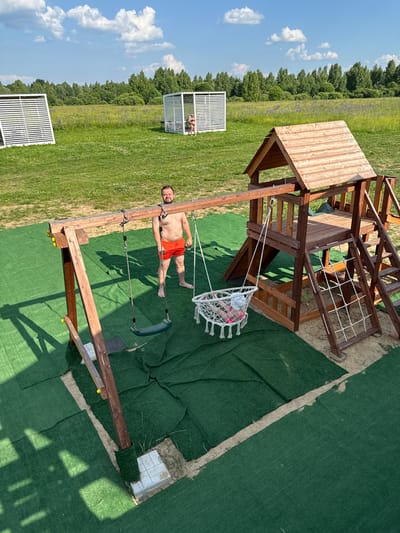 In the city of Koloksha, Russia, on a bright, sunny day, a man with a beard, wearing orange swim trunks, stands barefoot on a green artificial turf area. He is positioned near a wooden swing set, part of a larger wooden playground structure. The playground features a slide, a climbing wall with rope netting, and a wooden fort with a roof. A white macrame swing with a child inside is suspended from the swing set. To the left of the playground, another white structure with horizontal slats is visible, and further in the background, a second similar structure houses a person. The surrounding environment is a grassy field with scattered trees and a line of trees in the distance under a blue sky with white clouds. The ground beneath the playground is sand, and the artificial turf is laid out in sections.