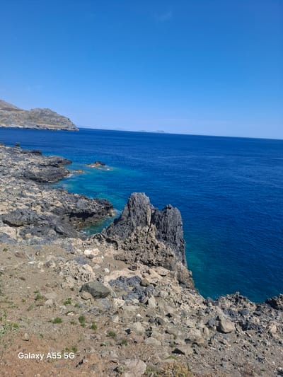 The image shows a coastal scene in Plakias, Greece. The camera angle is from a high vantage point, capturing a rocky coastline with deep blue water. Rocks are primarily grey and brown, varying in size and shape. The water is a deep blue color, transitioning to turquoise near the rocky edges where the water is shallow. A clear blue sky stretches above. In the lower right corner, there is a text that reads "Galaxy A55 5G" indicating the camera used to take the photo.