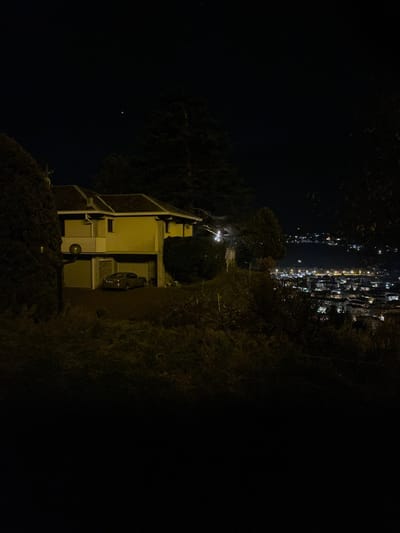 This nighttime photograph captures a serene elevated view overlooking a city in Sondrio, Italy. In the immediate foreground and midground, a multi-story building, likely residential, stands on the right side of the frame with light-colored walls and visible outdoor utility units. A vehicle is partially parked near the building. A paved path or driveway extends from the structure, flanked by dense, dark vegetation and trees that border the scene on both sides.
Beyond the immediate surroundings, a vast expanse of distant city lights shimmers horizontally across the upper portion of the image. These numerous artificial illuminations suggest a sprawling urban area nestled in a valley or across a wider plain, creating a striking contrast with the dark foreground. The night sky, a deep black, dominates the upper-left segment of the photograph, featuring a single, distinct bright star or planet.
The image conveys a quiet, tranquil atmosphere, highlighting the beauty of urban lights from a remote, elevated perspective. There are no people visible, and no specific activity is taking place. The scene emphasizes the natural darkness of the night interrupted by the glow of distant human habitation.