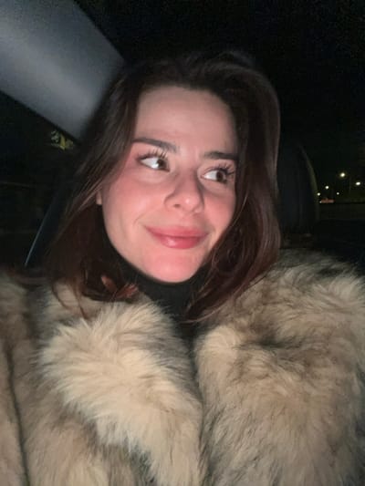 A woman with auburn hair and brown eyes smiles softly. She is facing left. The woman is wearing a black turtleneck and a large fur coat of varying shades of brown and tan. The background is mostly dark and out of focus, with some distant, blurry lights visible. She seems to be seated in a car with a headrest visible.
