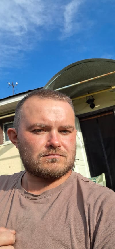 The media file is a close-up, selfie-style photograph of a man taken outdoors. The man has short, dark hair and a beard. He is wearing a plain, light brown t-shirt. His face is turned towards the camera, and he appears to be smiling slightly. His eyes are open, and his gaze is directed at the camera.
The background of the image is partially visible and suggests an outdoor setting. The sky is bright blue with some wispy white clouds, indicating it is daytime, likely sunny. There are also hints of a building or structure behind the man, with some architectural elements visible, possibly a house or a shed. A large, circular, metallic-looking object, possibly a lid or cover, is prominently featured in the upper left portion of the image, against the blue sky. Further back, a black, decorative lamp or sconce is visible on the wall of the structure. In the lower left, a utility pole with what appears to be lights or electrical components can be seen against the sky.
The lighting suggests it is daytime, with the sun casting shadows and highlighting the man's features. The overall mood appears casual and relaxed. No text is visible in the image. The location context provided is Verkhneural'sk, Russia.