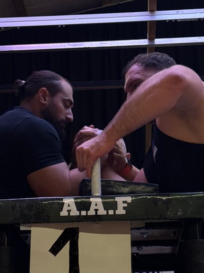 Two adult males are engaged in an arm-wrestling match indoors at a facility in Reisenberg, Austria. The male on the left has dark hair styled in a bun and a dark beard, wearing a black short-sleeved shirt. His right arm is positioned on the arm wrestling table's pad. The male on the right has short dark hair, wears a dark sleeveless top, and has a red wristband on his left wrist. His left arm is similarly positioned on the table's pad. Their hands are clasped in the center. A third, unidentified hand, likely an official's, is positioned over their clasped hands, holding a light-colored vertical peg on the table. The arm wrestling table features dark-colored padding and displays the white letters "A.A.F" on its front edge. Below the table, a white sign with a large black numeral "1" is visible. The background is dark, showing metallic overhead structural beams.