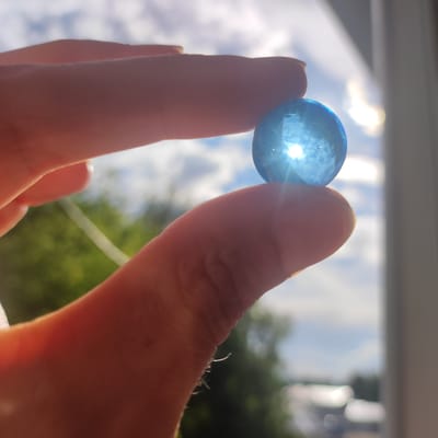 In this close-up, an individual's fingers gently hold a translucent blue marble between the thumb and forefinger. The marble is illuminated from behind by bright sunlight, causing it to glow and refract the light into a starburst pattern. The background is softly blurred, showing a daytime sky with scattered white clouds and hints of green foliage, suggesting an outdoor setting. The lighting indicates it is likely daytime, possibly late morning or early afternoon, with bright sunshine. The overall mood is calm and contemplative, with a focus on the simple beauty of light interacting with the marble. No text is visible in the image.