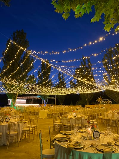 The image depicts an outdoor wedding reception setup at dusk in Bullas, Spain. Numerous round tables, draped in white tablecloths and set with gold chairs, are arranged in rows. Each table is meticulously set with silverware, glassware, plates, napkins, and decorative centerpieces featuring candles and flowers. The scene is illuminated by an extensive network of string lights that are draped across the trees and over the dining area, creating a warm and festive ambiance against the deep blue twilight sky. In the background, more tables and chairs extend into the distance, suggesting a large event. The surrounding environment includes trees with lush green foliage and what appears to be a rustic stone wall in the far background. The overall impression is one of an elegant and atmospheric evening celebration.