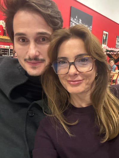The image depicts two individuals in the foreground of an indoor public establishment in Décines-Charpieu, France. The scene captures an adult male partially visible on the left, and an adult female prominently featured on the right.
The male has dark, wavy hair, dark eyes, and a dark mustache. He wears a dark turtleneck beneath a dark, possibly charcoal-colored, heavy coat or jacket. His expression is a slight smile directed towards the camera.
The female has long, light brown hair, parted on the side, and wears large, square-framed eyeglasses with a tortoiseshell pattern. She has a small gold-colored hoop earring visible in her right ear. Her facial expression is a gentle smile, also directed at the camera. She is wearing a dark purple or maroon long-sleeved top. Their close proximity suggests they are posing together for a photograph, likely a selfie.
The background features a predominantly bright red wall. Decorative elements on the wall include a large, rectangular framed picture depicting a night cityscape (likely New York City) positioned above and behind the female's head. To the right of this, a smaller framed image is partially visible. Below the cityscape and to the right of the female, a decorative arrangement resembling pink and white cherry blossoms is present, partially obscuring a dark panel or partition. To the far right, a young individual, possibly a child or teenager with light hair, is visible seated at a table, out of focus. The background environment suggests a casual dining establishment with tables and chairs.
