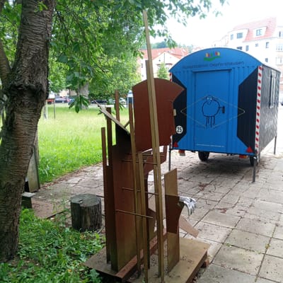 A rusted metal sculpture stands in front of a blue trailer with the words "Kunststation Oepfershausen" printed on it, in Eisenach, Germany. The trailer has a drawing of a sheep on the back and the number "25" is visible on a round sign. A tree trunk is to the left of the sculpture, and a grassy area with trees is in the background. The ground is paved with concrete tiles. A white glove is resting on one of the metal parts of the sculpture.