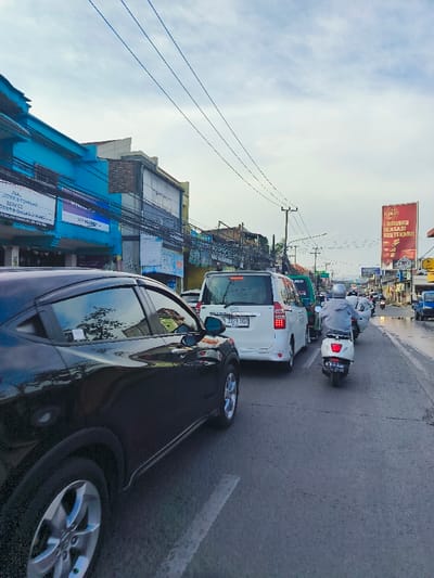 The image shows a street scene in Bojongsoang, Indonesia, located at the coordinates -6.97 latitude and 107.637 longitude. The scene is a busy street with a line of vehicles stopped in traffic. There are several cars, including a black SUV, a white minivan, and a blue van. In front of the cars is a person riding a white motorcycle. The person is wearing a grey jacket and black helmet. There are buildings on both sides of the street, and power lines run overhead. The sky is cloudy, and there is a large billboard visible in the background. The street is paved with asphalt and has white lane markings. The overall scene is typical of a busy urban area.