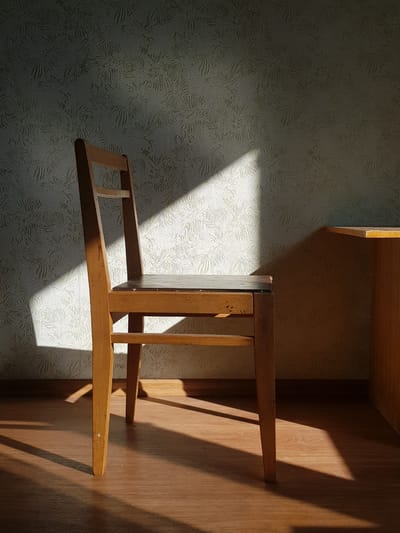 An empty wooden chair sits in the foreground, bathed in a strong diagonal shaft of sunlight that cuts across the wall and floor. The wall behind the chair has a textured, repeating pattern of light and shadow. To the right of the chair is the corner of a wooden desk or table. The floor is made of wooden planks. The scene appears to be indoors, possibly in an apartment or office. There is no indication of Kurchatov, Kazakhstan.