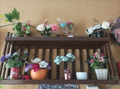 The image displays an interior scene in Pasir Muncang, Indonesia, featuring a two-tiered dark brown wooden shelf mounted against a textured, light brown or beige wall.
On the upper shelf, from left to right: a clear glass vase holds artificial green ivy and small pink flowers; a second clear glass vase contains artificial white and pink roses; two separate bouquets of artificial blue and red flowers, each wrapped in clear plastic with a red ribbon, stand upright; and a black pot holds artificial white and cream-colored roses. A large, dark-colored artwork or tapestry with floral patterns is partially visible on the far right.
On the lower shelf, from left to right: a red can, decorated with a "Cars" character (Lightning McQueen), serves as a planter for artificial purple flowers and green foliage; a small red ladybug-shaped object rests beside it; an orange ceramic pot contains artificial white, purple, and red flowers; a pink glass vase holds artificial light blue or teal succulent-like flowers on wire stems; a small white ceramic bowl is present; and a light-colored woven basket-like pot contains artificial pink and green flowers. A green artificial vine drapes from the right side of the lower shelf. Below the bottom shelf, a dark fabric with a light pattern is partially visible, along with a white object on the bottom right. No individuals or active interactions are present in the scene.
