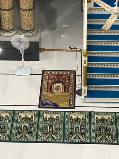 A room features a decorative ceiling with blue and white stripes and gold trim, under which hangs a white ceiling fan. A brown and gold carpet hangs on the wall next to a green prayer rug with repeated geometric patterns. Below, a pedestal supporting a microphone is visible, along with a free-standing fan and decorative pillars with a patterned surface. The wall is white with thin, vertical black lines.