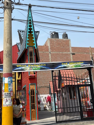 Here is a detailed description of the image:
**Scene:** A street scene in Aucallama, Peru, with a focus on the facade of a vibrantly colored building.
**Subjects:**
* **Building:** The building is the main subject, notable for its eclectic architecture, including an ornate, multi-layered roof with turquoise accents, red and yellow striped walls, and arched windows.
* **Woman:** A woman is visible on the right side of the frame. She is wearing sunglasses, a tan jacket, and black pants. She appears to be standing, partially obscured by a yellow post and some fencing.
* **Advertising Banner:** There is a tall, vertical banner attached to the building and a nearby post. It reads "Mas Delgada del Mundo" (Slimmer World) and "Entrada" (Entrance) with a visual element suggesting a globe.
**Objects:**
* **Utility Pole:** A prominent concrete utility pole is in the foreground, covered with various posters and stickers.
* **Water Tanks:** Two large, black water tanks are visible on the roof of a brick building adjacent to the main subject building.
* **Fencing:** Black metal fencing is present on the right side of the frame, partially obscuring the building behind it.
* **Posters/Stickers:** The utility pole is plastered with advertising posters, including some mentioning a lawyer named Gaby Sota and selling land.
**Actions/Interactions:**
* The woman appears to be standing and possibly looking at something inside the building or in the area.
**Environment:**
* **Location:** Aucallama, Peru.
* **Weather:** The sky is clear and blue, suggesting a sunny day.
* **Background:** The background includes a brick building under construction and numerous utility wires.
**Forensic Details:**
* The image is taken at an angle, with a slight upward tilt.
* The utility pole is positioned in the foreground, partially obscuring the building.
* The vibrant colors and architectural details of the building are prominent.
* The state of the utility pole and surrounding buildings indicates a somewhat urban or semi-urban environment.
* The advertising banner is positioned to attract attention to the building.
* The building appears to be old, as it is located in the small city of Aucallama in Peru.
This description provides a clear, forensic overview of the scene as depicted in the image.