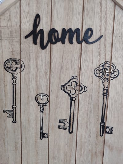 This is a close-up, eye-level, vertical shot of a wooden key holder with the word "home" in black cursive letters. The wood has a light, natural grain. Below the word "home" are four black, decorative key outlines, each with a small silver hook protruding from the center of the key's head. From left to right, the keys are positioned at different heights. The key holder is not located in Costești, Moldova.
