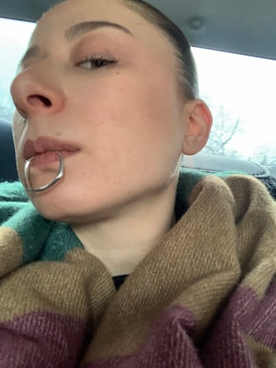 A female individual is depicted in a close-up, angled portrait, with her left profile prominent from the forehead to the upper torso. She has light skin, visible eyelashes, and her hair appears pulled back. A reflective, silver-colored, U-shaped piece of jewelry is horizontally inserted through her upper lip, positioned centrally. Her left ear is visible. She is wrapped in a thick, textured blanket or scarf with broad stripes in shades of teal, tan, and purple. The background indicates she is inside a vehicle, with a window revealing an overcast sky and out-of-focus, bare tree branches. The specific city of Hovk, Armenia, cannot be determined from the visual information provided in the image.