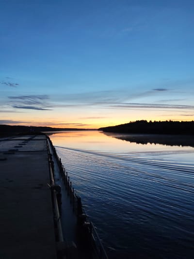 The image captures a serene twilight scene over a body of water, likely a river or canal, in Andriushino, Russia. The sky is a gradient of deep blue at the top, transitioning to lighter blues and wisps of pink and orange near the horizon, indicative of a sunset or sunrise. A dark, silhouetted treeline forms the distant bank of the water. In the foreground, a concrete structure, possibly a dock or pier, extends into the water. A metal railing runs along the edge of this structure, separating it from the water. The surface of the water is rippled, with a distinct wake pattern suggesting movement, perhaps from a passing boat. The overall atmosphere is calm and tranquil. There are no people visible in the frame.