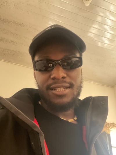 A close-up, low-angle selfie shows a Black man wearing a black baseball cap, black sunglasses, and a black jacket. He has a light beard and is wearing a beaded necklace. The background is a ceiling with wooden panels and a light fixture, and a wall that appears to be painted yellow or cream. The image is lit from above and slightly to the right. The man is looking directly at the camera with his mouth slightly open, showing his teeth. The reflection in his sunglasses is visible but blurry. The scene appears to be indoors. There is no indication of the location being Rahwol Chom, Nigeria.