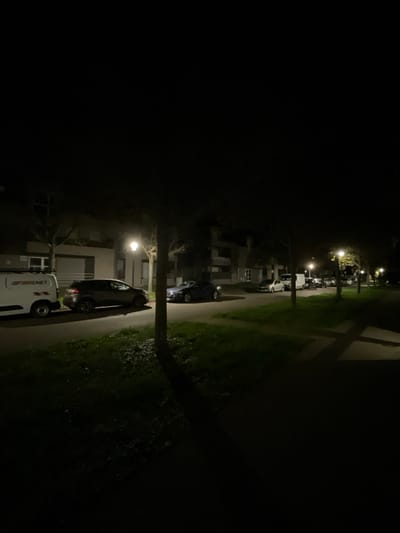 A dimly lit night scene shows a street with parked cars along the right side. A sidewalk runs adjacent to a grassy area. Streetlights cast a soft glow. Buildings are visible behind the vehicles. The left side of the image is predominantly dark. Trees are present, adding to the shadowy atmosphere.
