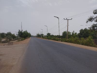 The image shows a paved road lined with solar powered streetlights and power lines in Kwadon, Nigeria. There are trees on both sides of the road, along with some dirt patches, suggesting a rural setting. The road is empty of vehicles and people, and the overall atmosphere is calm and quiet. The sky is a pale gray, suggesting a cloudy day.