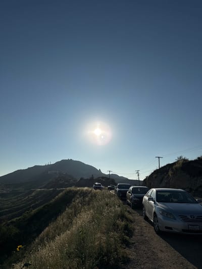 This image depicts a clear, sunny outdoor scene in the Unincorporated Santa Monica Mountains, United States of America. The sun is prominent and bright in the upper center of the clear blue sky, casting a strong backlight. A line of approximately five stationary passenger vehicles is visible on the right side of a dirt or gravel road, heading into the distance. The closest vehicle is a silver Toyota Camry sedan with the license plate "6VVV457". Utility poles with power lines run alongside the right edge of the road. To the left, a large, dark mountain with multiple communication towers or structures on its peak dominates the mid-ground. Rolling hills covered in dry, golden grasses occupy the left foreground and mid-ground. A single yellow sunflower is visible in the bottom left corner of the foreground. No individuals are visible.