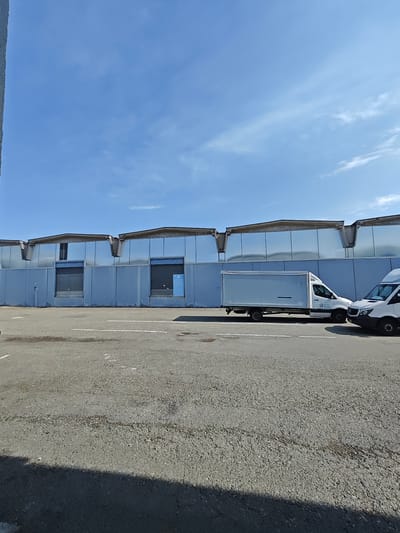In Settimo Torinese, Italy, a grey asphalt lot sits before a long, pale blue industrial building under a bright, clear blue sky with scattered white clouds. The building features reflective silver panels across its upper facade and large, closed grey roller doors on its lower level. Two white cargo vans are parked in the lot. The van on the left is a box truck with a long, white cargo area and the logo "ICE" and "ACQUA FRESCA" visible on its side. The van on the right is a smaller, standard cargo van. The lot has faint white painted lines, suggesting parking spaces. The ground is weathered and shows some patches of oil stains.