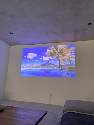 A wall-mounted television displays a tropical beach scene with palm trees, a blue sky, and mountains in the background. The TV is situated between a textured gray wall on the left and a white wall. A couch partially appears on the right, and a wooden table sits below the couch, with visible power cords. An electrical outlet is visible on the white wall near the table.