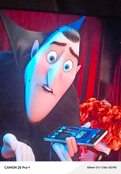 The image is a close-up shot of a cartoon character, possibly Dracula from the Hotel Transylvania franchise, with bright blue lighting illuminating his face from the left and pinkish-red lighting from the right. His eyes are wide open, and he appears to be looking at something off-camera. He is holding a smartphone in his hands. The smartphone screen displays the time "3:08" in glowing blue digits, with other interface elements also visible. Behind the character, a red, striped curtain or backdrop is visible, and to the right, a cluster of red, flower-like objects. The bottom of the image contains text: "CAMON 20 Pro" with a red dot, and "150mm f/1.7 1/50s ISO792". There is no discernible information within the image that suggests a location in the city of Вышков, Russia.