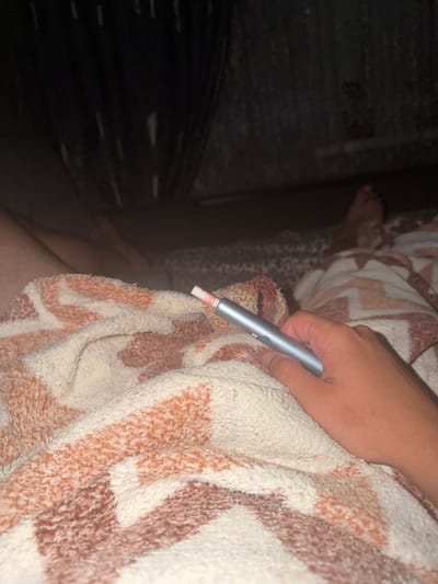 The image shows a close-up of a hand holding a silver e-cigarette device over a patterned blanket. The blanket is cream-colored with a pattern of rust-colored geometric shapes. In the background, a person's feet are visible, also covered by a blanket. The environment appears to be indoors, possibly a bedroom. There is no indication of the specific location within the city of Avdarma, Moldova.