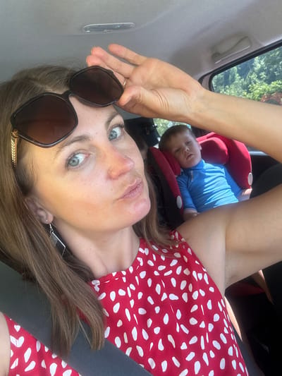 A woman is taking a selfie in a car, with a child in a car seat in the background. The woman is wearing sunglasses and a red and white polka dot dress. The child is wearing a blue shirt and appears to be sleeping. The car appears to be in a rural or suburban area, with trees visible outside the window. The location is not specified in the image, but the prompt suggests it could be in Lavans-lès-Saint-Claude, France.