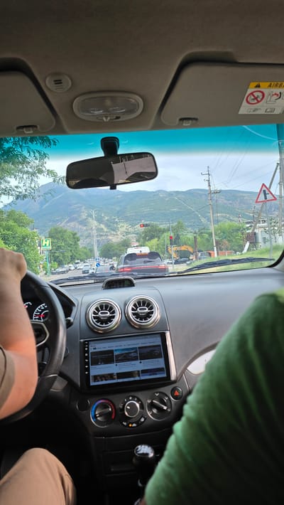 The image is taken from the interior of a car, looking forward through the windshield. The driver's hand is visible on the steering wheel on the left. The dashboard features an infotainment screen displaying what appears to be a YouTube interface. There are two circular air vents above the screen.
Outside the car, the scene depicts a road with moderate traffic, including cars, a truck, and an excavator. The road is lined with trees and buildings. In the background, a large mountain range is visible under a cloudy sky. A traffic light is showing red. A triangular road sign indicating a yield or a curve is also present on the right side of the road. The overall impression is a view of a city or town street with a scenic mountainous backdrop. The presence of the road signs and the general streetscape suggests an urban or semi-urban environment. The text on the screen of the infotainment system and the style of the road signs are consistent with the location of Charvak, Uzbekistan.