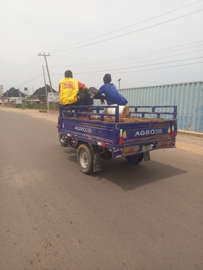This media file captures a scene in Baccure Kofare, Nigeria, on an overcast day. The main focus is a blue, three-wheeled vehicle, likely a utility cart, loaded with soil and a white bucket. Two individuals are seated in the back of the cart. One, wearing a yellow shirt with red accents and possibly a logo that reads "Roasted," is seated towards the front. The other, in a blue shirt, sits behind him.

The cart itself has the text "AGRO200" prominently displayed on its side. The license plate, partially visible, reads "NT-502VE". The vehicle is on a paved road, with a sandy or dirt shoulder. In the background, there are buildings with dark, sloped roofs, suggesting a residential or commercial area. Utility poles with power lines crisscross the sky, and a large, corrugated metal structure, possibly a shipping container or industrial building, lines the right side of the frame.

The overall impression is of a practical, everyday scene, possibly related to agriculture or construction, given the load and the "AGRO200" text. The people appear to be engaged in transport, with no strong emotional expressions discernible from their postures. The lighting suggests it's daytime, but the cloudy conditions create a subdued atmosphere.