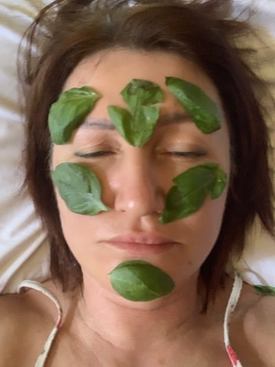 The image shows a person lying down with their eyes closed, resting on a white surface. Their face is adorned with several fresh green leaves, strategically placed across their forehead, cheeks, and chin. The leaves appear to be basil or a similar herb, suggesting a natural skincare or relaxation ritual. The person has dark hair that frames their face and extends to the sides. They are wearing a light-colored garment, possibly a camisole or dress, with a thin strap visible on the right side of the frame. The lighting is soft and diffused, creating a serene atmosphere. The overall impression is one of peacefulness and self-care. The context of San Quirico d'Orcia, Italy, suggests a possible connection to the region's agricultural abundance or a tranquil rural setting.